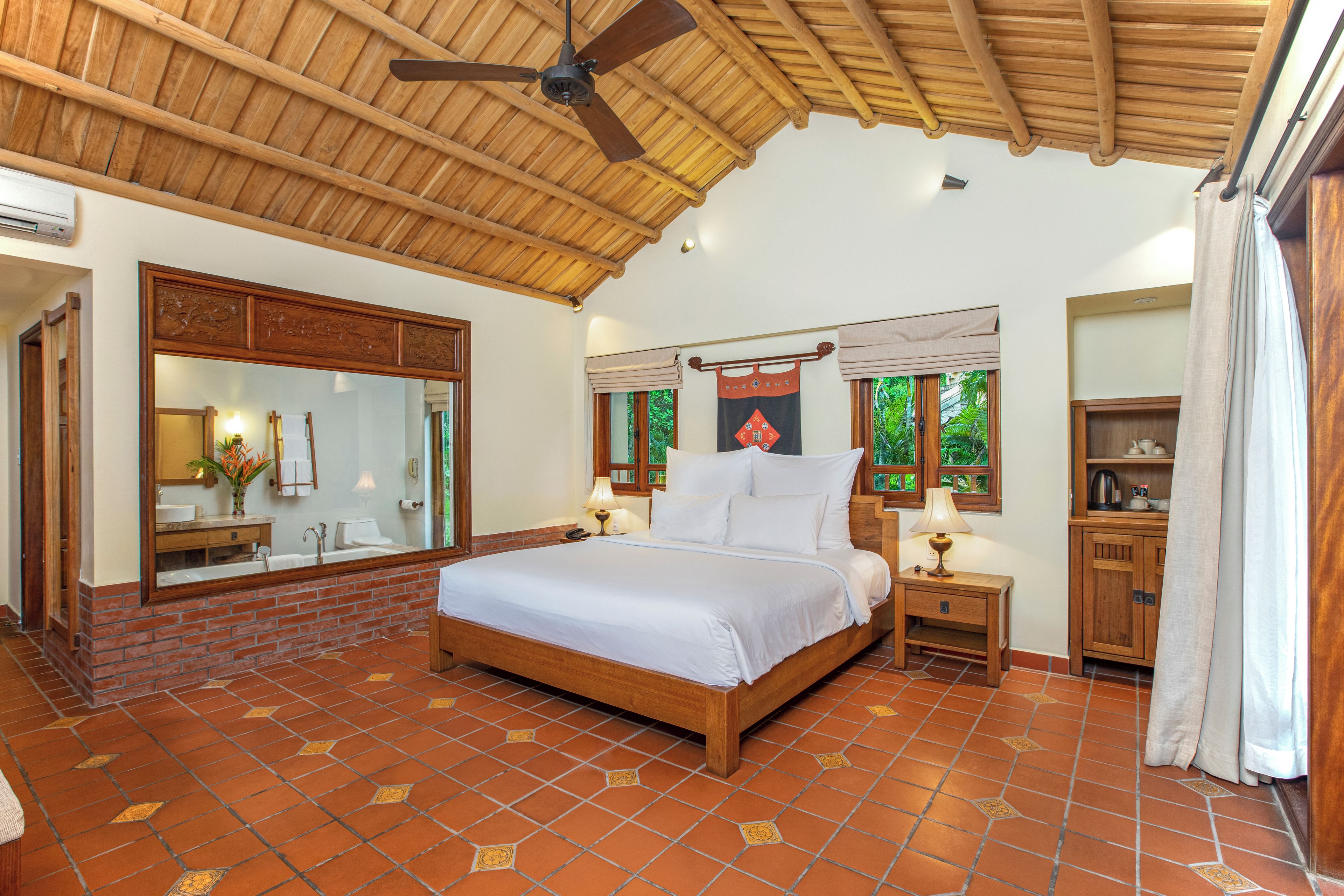 A spacious bedroom with a large bed, a wooden ceiling fan, a brick wall, and a tiled floor.