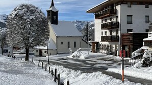 Una calle nevada con una iglesia, un edificio moderno con balcones y una montaña al fondo.