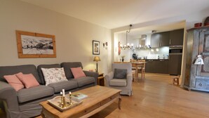 Una sala de estar con sofá, sillón y mesa de centro. Se observa una cocina al fondo con una mesa de comedor y sillas.