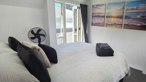 Ein Hotelzimmer mit einem Bett, einem Standventilator, einer Handtuchauslage und einem großen Fenster mit Blick auf den Strand.