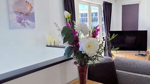 Ein modernes Wohnzimmer mit einem Holzesstisch, einem Blumenstrauß, einem Flachbildfernseher und einem großen Fenster mit Vorhängen.