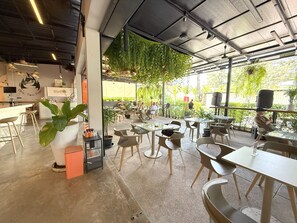 Un café moderno con mesas dentro y fuera, una zona de bar y plantas colgantes.