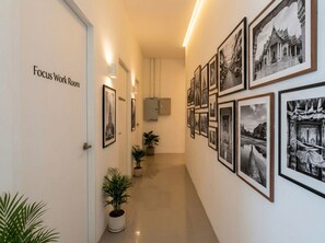 A hallway with framed black and white photographs on the walls, a sign for 'Focus Work Room,' and potted plants along the corridor.