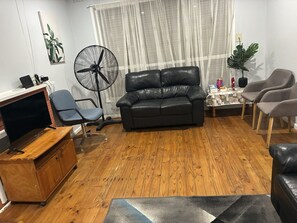 Een woonkamer met een zwarte leren bank, een houten kast, een televisie, een ventilator en een salontafel.