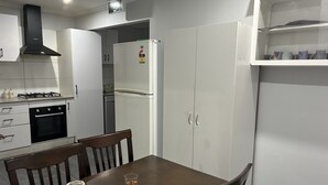 A compact kitchen with white cabinets, a stove, and a refrigerator. There is a dining table with chairs and a glass of water. - Three furnished bedrooms and two bathrooms house (Deer park)