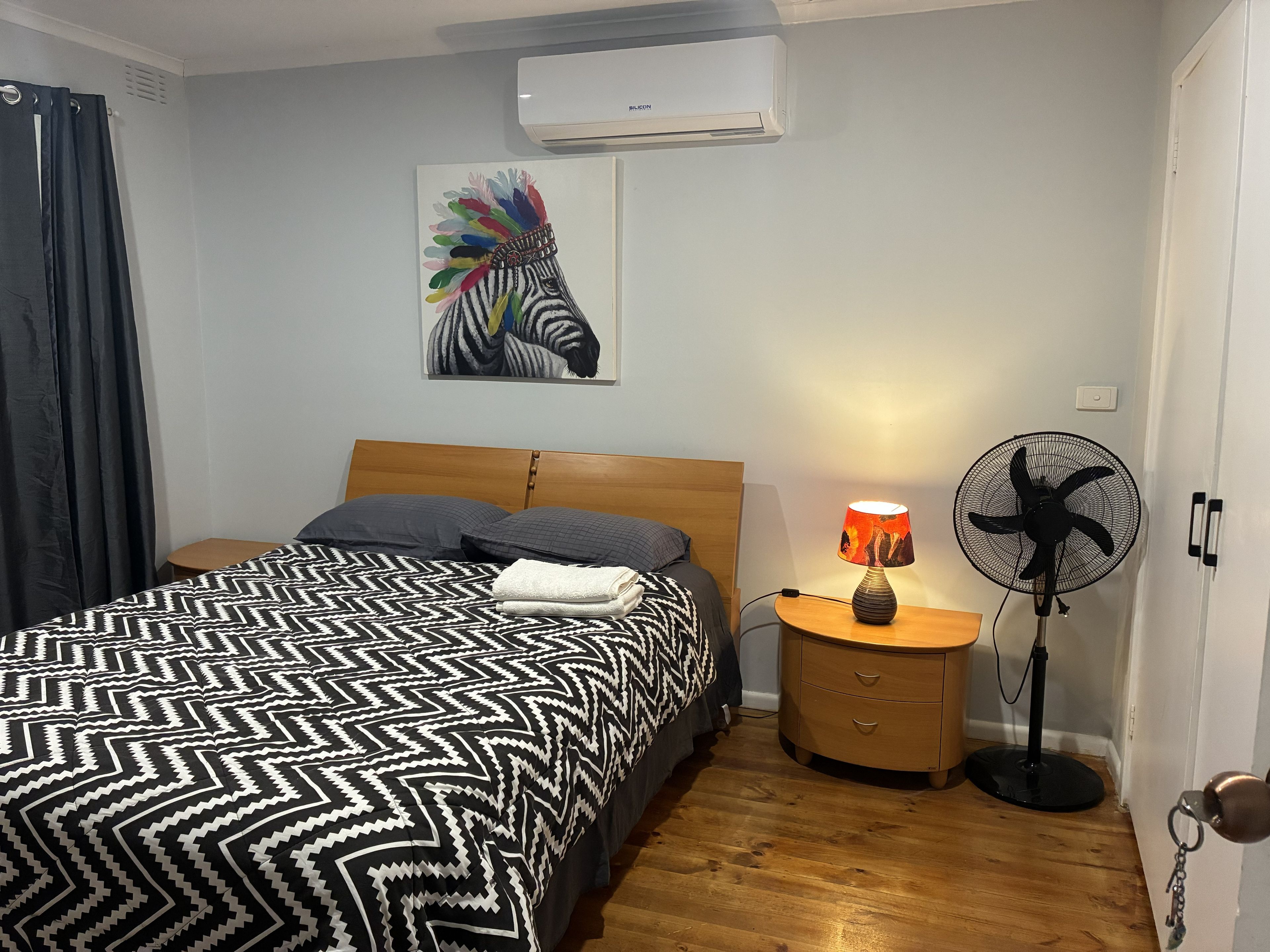 Un dormitorio con cama, mesita de noche, lámpara, ventilador y aire acondicionado instalado en la pared.