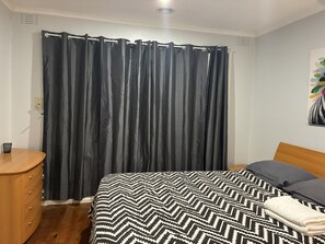 Une chambre à coucher avec un lit, une table de chevet en bois, une couvre-lit à motifs noir et blanc, un rideau noir et un luminaire fixé au mur.