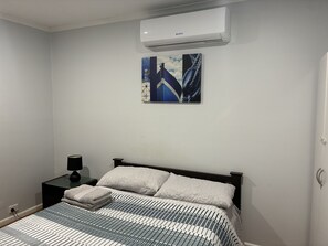 A bedroom with a bed, a nightstand, a lamp, an air conditioning unit, and a wall-mounted artwork.