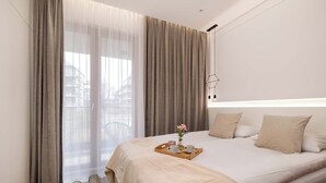 Hotellihuone, jossa on suuri sänky, beigenväriset verhot, tarjotinalus kuppeineen ja kukkineen sekä näkymä ulkona oleviin rakennuksiin.