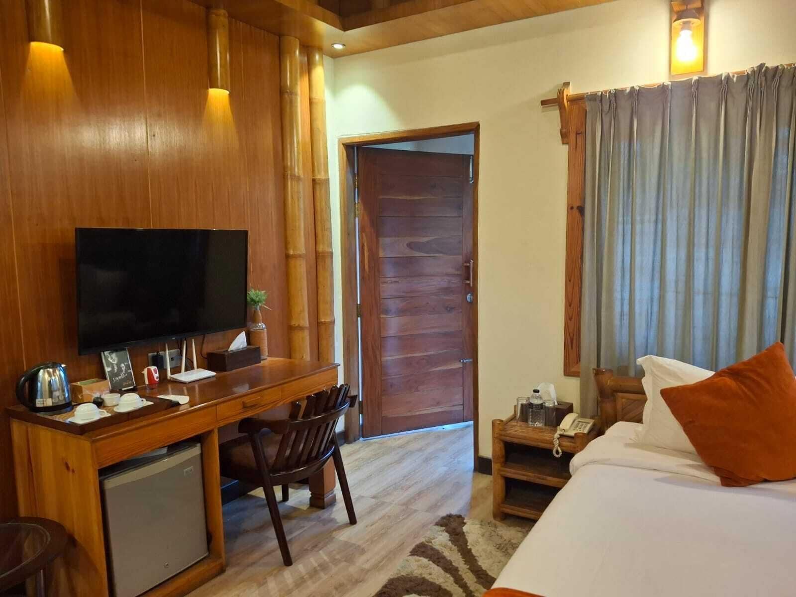 A hotel room with a wooden desk, television, bed, and a small bedside table with a lamp.