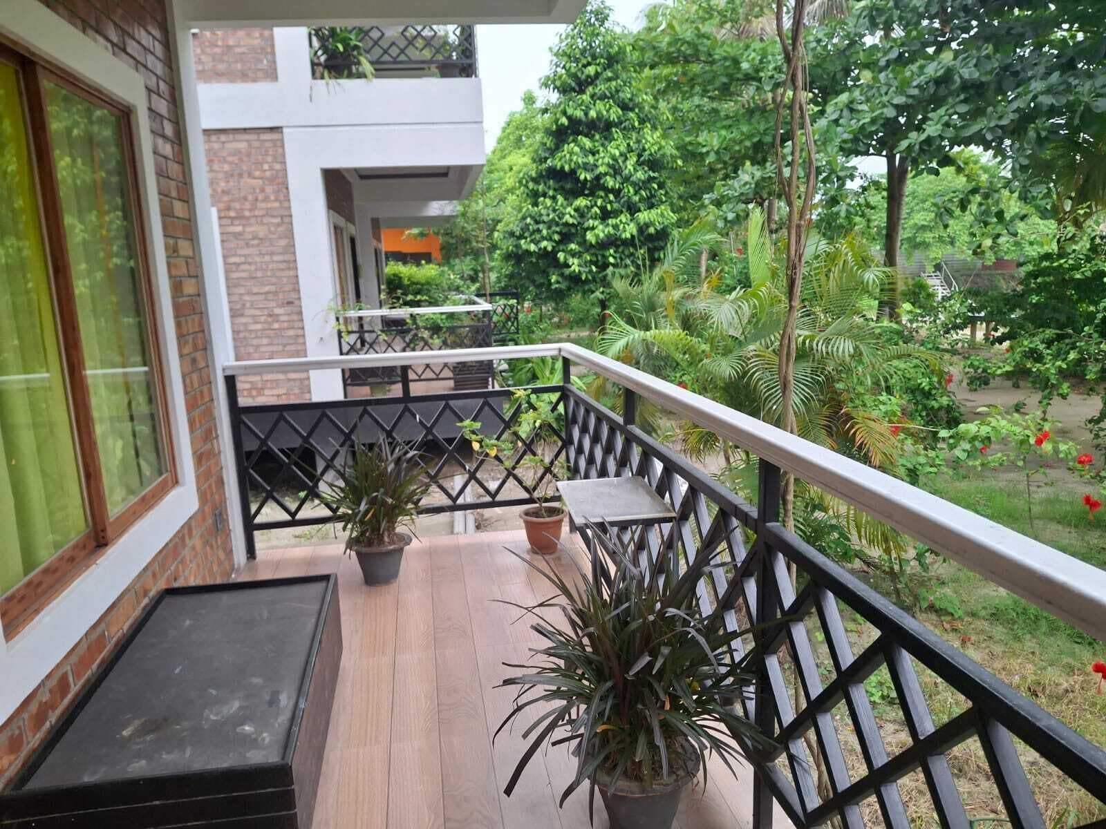 A balcony with potted plants, a wooden floor, and a view of a garden with trees and a building in the background.