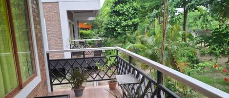 A balcony with potted plants, a wooden floor, and a view of a garden with trees and a building in the background.