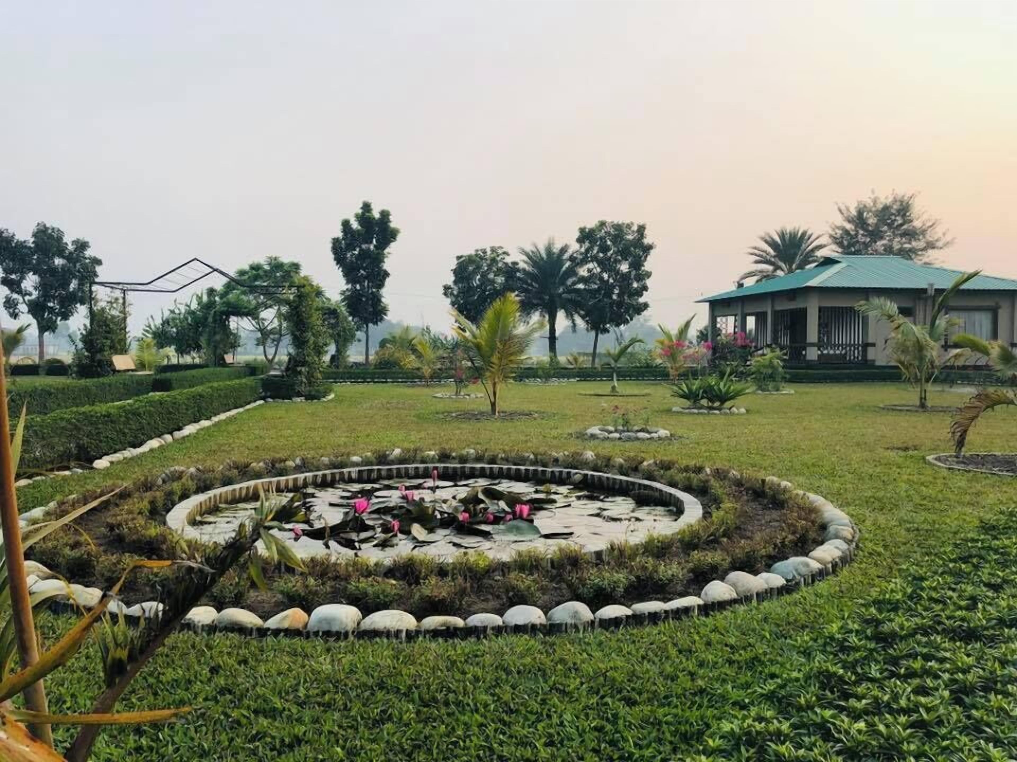 Um jardim bem cuidado com um lago circular, sebes aparadas e um edifício com telhado verde ao fundo.