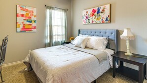 A bedroom with a bed, a nightstand, a lamp, a window with curtains, and two wall art pieces.