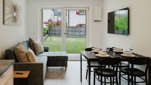 Ein modernes Wohnzimmer mit einer Couch, einem Esstisch und Stühlen. Ein großes Schiebeglasfenster führt in einen Garten mit Zaun und viel Grün.