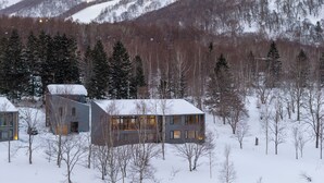 A modern building with a snow-covered roof, surrounded by trees and a mountainous backdrop. - Mokuren by H2 Life (Niseko)