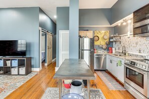 Una cocina moderna con un refrigerador, horno y microondas de acero inoxidable. Hay una mesa de comedor de madera con cubierta metálica y un televisor sobre un soporte.