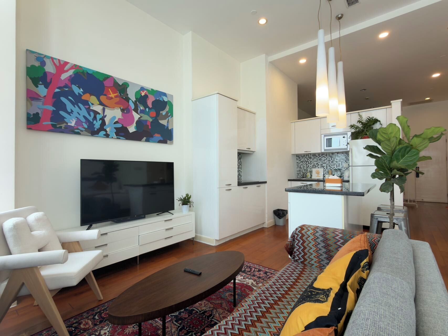 Una sala de estar moderna con televisor de pantalla plana, muebles blancos, un cuadro abstracto de colores, un tapete estampado y una cocina con armarios blancos y un microondas.