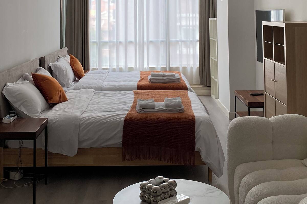 A hotel room with a bed, bedside tables, a TV, a wardrobe, and a view of the city through sheer curtains.