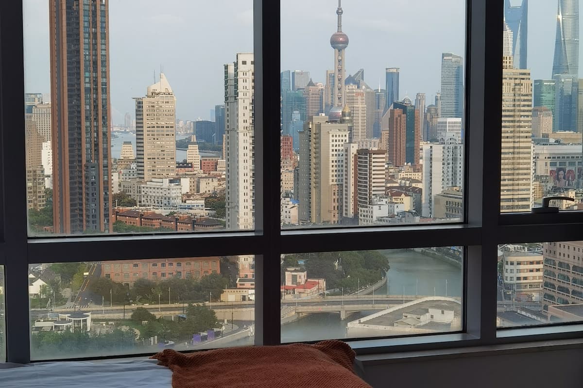 A hotel room with a large window offering a city skyline view, a neatly made bed, and a red throw blanket.