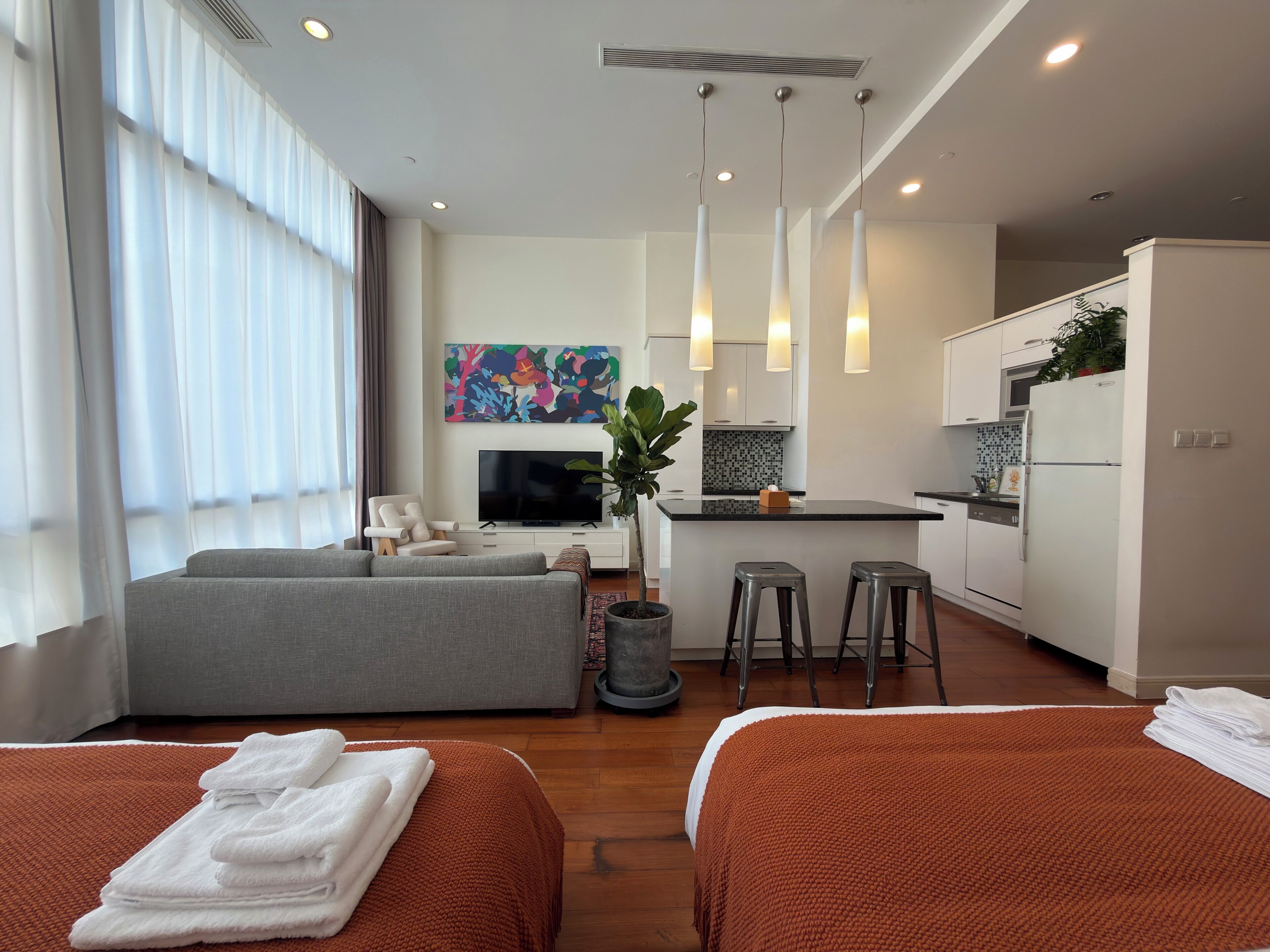 A modern hotel room with a sofa, two beds, a TV, a kitchen area, and a large window with curtains.