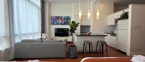 A modern hotel room with a sofa, two beds, a TV, a kitchen area, and a large window with curtains.