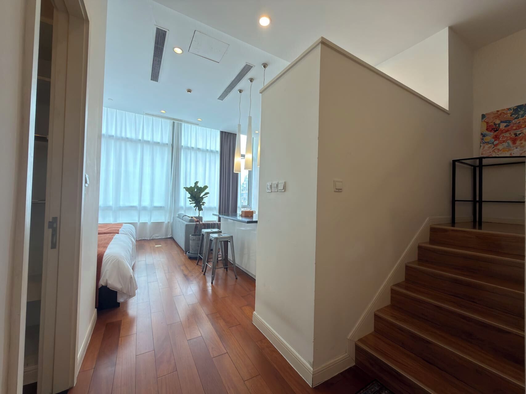 A hotel room with a wooden floor, a bed, a desk with a chair, a plant, and a staircase.