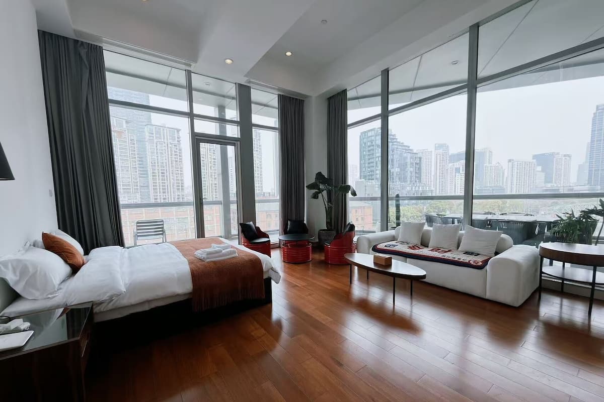 A modern hotel room with a bed, sofa, coffee table, and large windows offering a city view.
