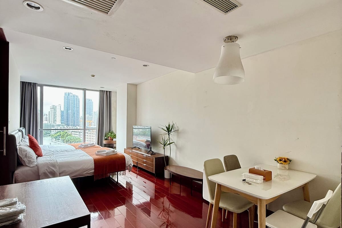 A modern hotel room with a bed, a dining area with a table and chairs, a TV, and a large window offering a city view.