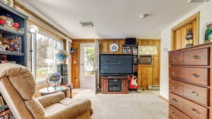 A living room with a beige sofa, a television on a wooden stand, a wooden cabinet, and a bookshelf filled with books and decorative items. - Hike & Explore Western Mass: Peaceful Studio (Palmer)