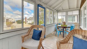 A covered porch with wicker chairs and a table, large windows, and a view of the neighborhood. - Steps to Beach: Timeless Lower Cape Cottage (Brewster)