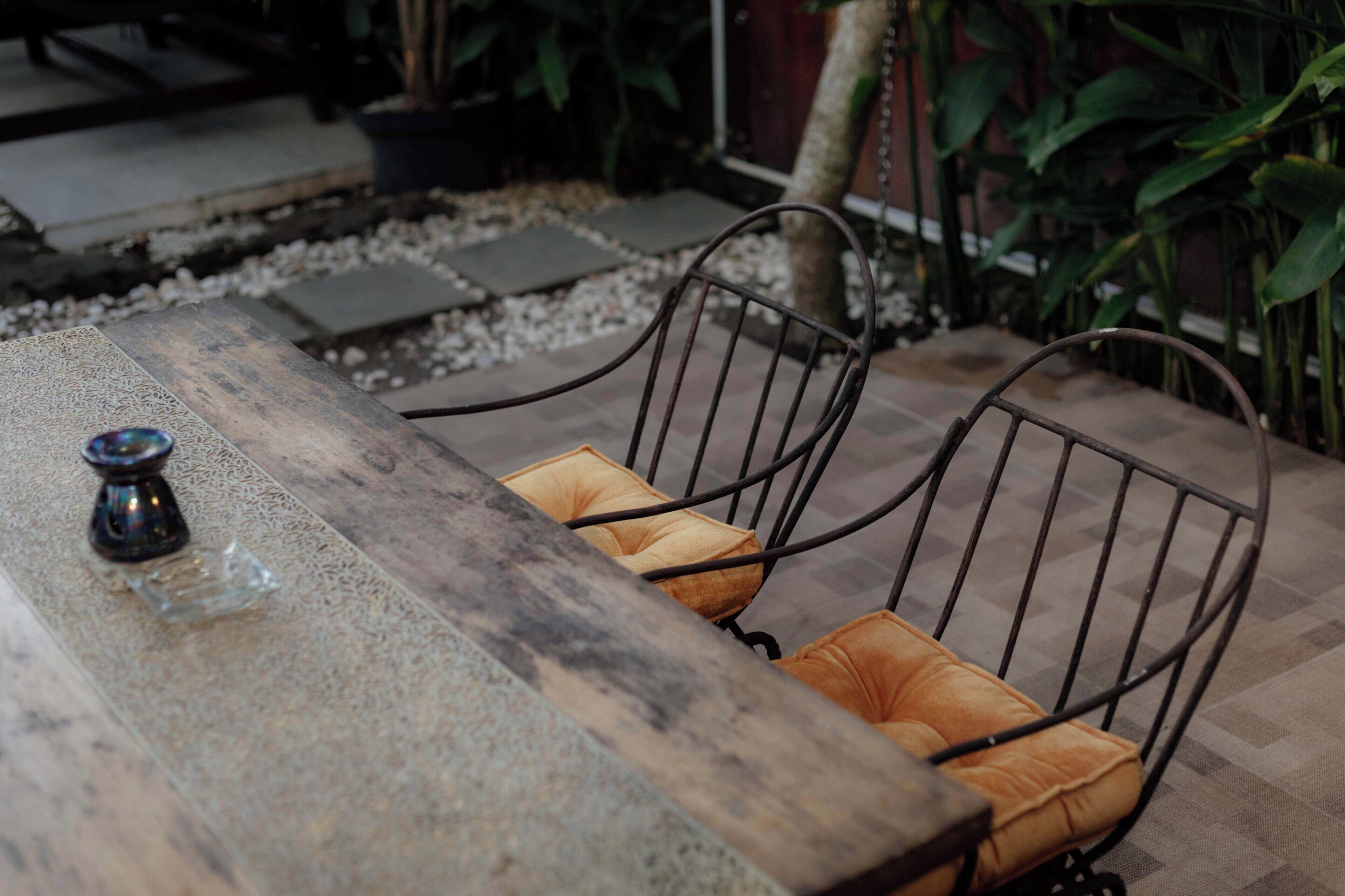 Une table en bois, des chaises en métal et des coussins, un objet décoratif en verre, et un espace jardin agrémenté de plantes et de pas japonais.