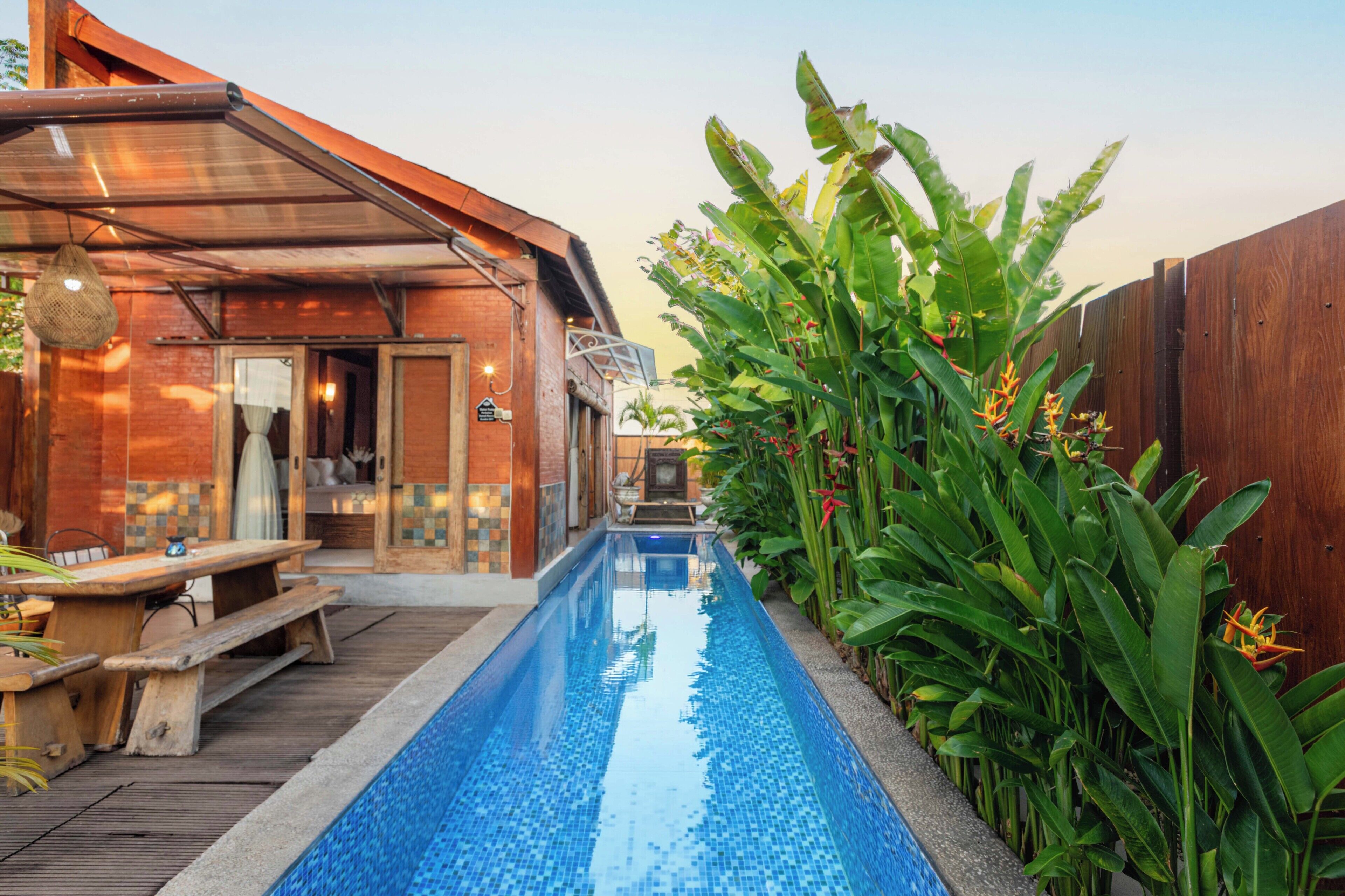 A pool area with a wooden deck, a covered seating area, and lush green plants.