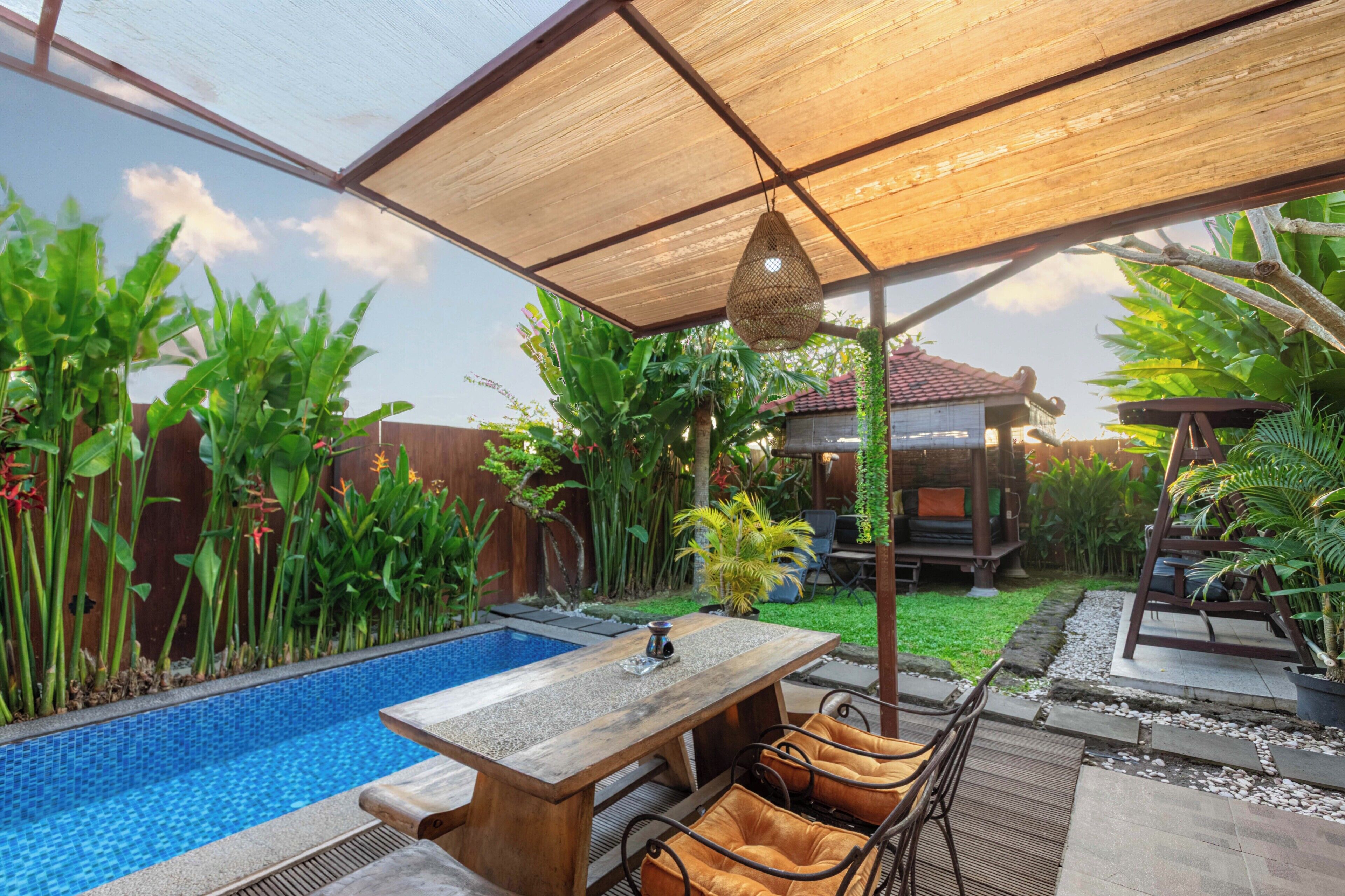 Un área junto a la piscina con mesa y sillas de madera, rodeada de exuberante vegetación y un pequeño pabellón.