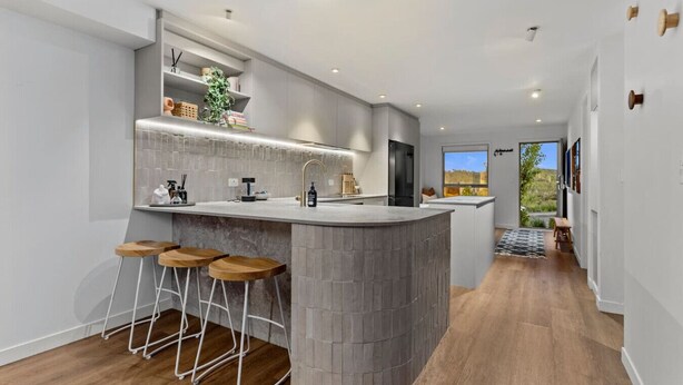 A modern kitchen with a central island, bar stools, and a view to the outside. - Spacious Coastal Luxe Home with Sea View & Parking (Auckland Hibiscus Coast)