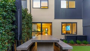 A modern house with a large patio table, a wooden deck, and a well-maintained garden. - Spacious Coastal Luxe Home with Sea View & Parking (Auckland Hibiscus Coast)