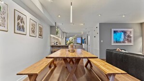 A spacious room with a long wooden table, a sofa, and framed artwork on the walls. - Spacious Coastal Luxe Home with Sea View & Parking (Auckland Hibiscus Coast)