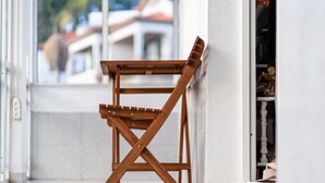 Ein zusammenklappbarer Holzhstuhl vor einer weißen Wand mit einem Fenster, durch das ein Gebäude zu sehen ist.