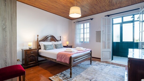 Ein Schlafzimmer mit Holzbalkendecke, einem Bett mit Kopfteil, Nachttischen, einem roten Sessel und Blick durch eine offene Tür.