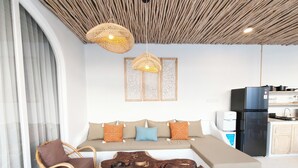 A modern living room with a sofa, a wicker chair, a coffee table, and a kitchen area with a refrigerator and a water dispenser. - Villa Vinodayan Uluwatu (Ungasan)