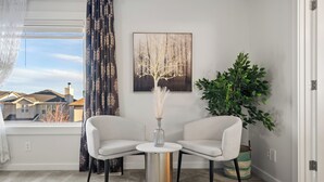 Een moderne woonkamer met twee witte fauteuils, een klein rond tafeltje, een vaas met gedroogde planten, een ingelijst schilderij aan de muur, en een grote groene plant.