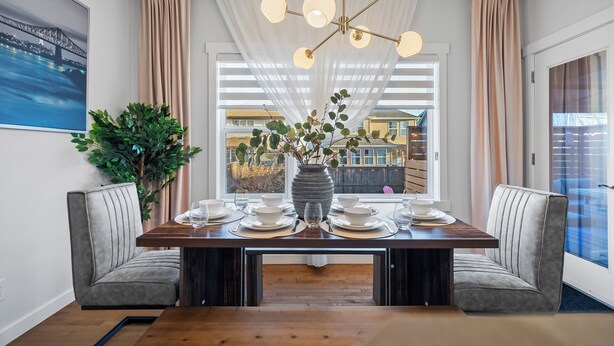 Een eetgedeelte met een houten tafel voor vier personen, omringd door twee grijze gestoffeerde stoelen. Een groot raam met witte zonwering laat natuurlijk licht binnen, en er is een stadsgezicht te zien door het raam.