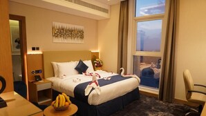 A hotel room with a bed, a desk, a chair, a small table with fruit, and a window with a view of the city. - Al Hayat Hotel (Sharjah)