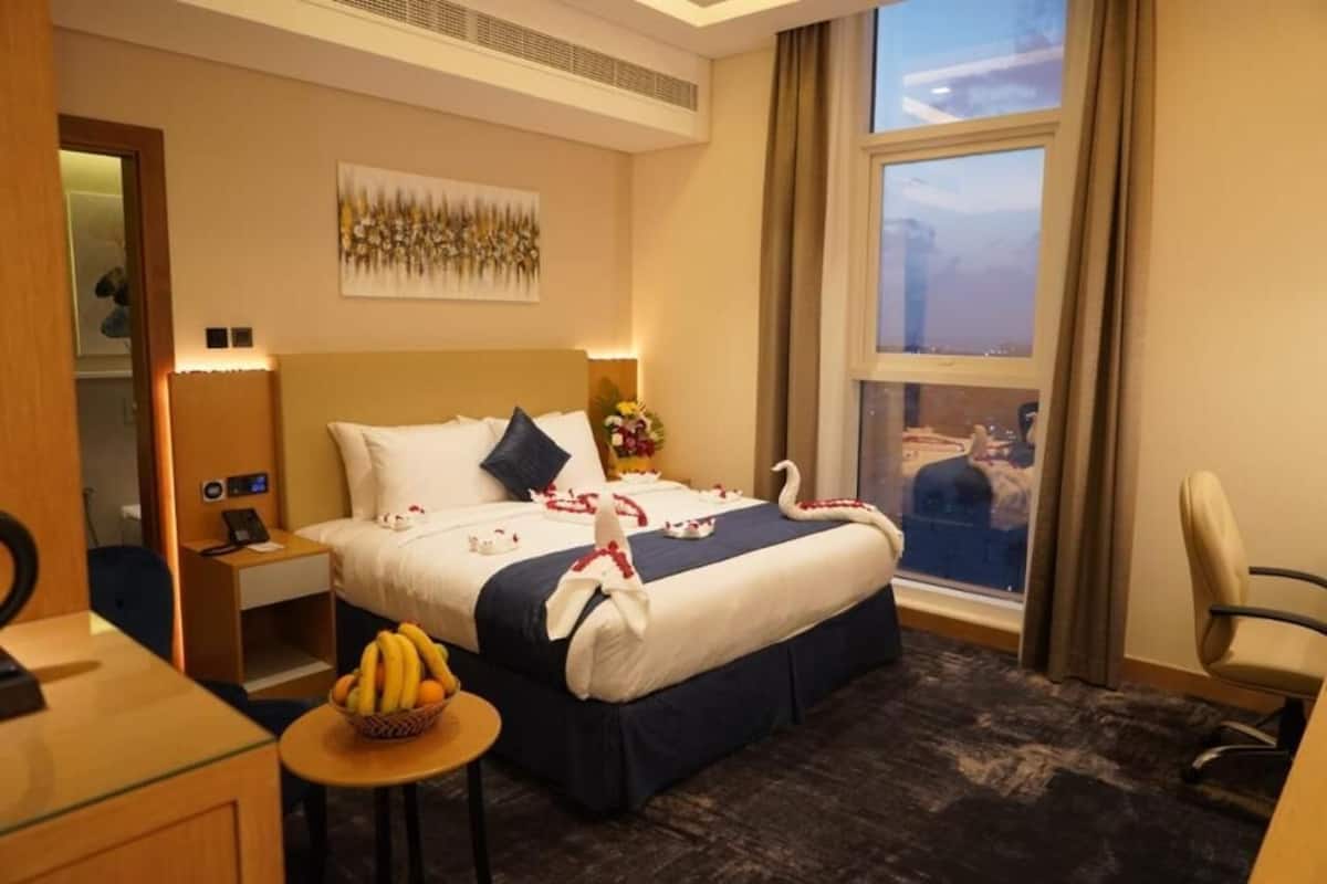 A hotel room with a bed, a desk, a chair, a small table with fruit, and a window with a view of the city.