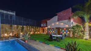 A swimming pool with a wooden deck, a patio with a table and chairs, and a building with colorful window decorations. - Nimbal Dhani (Jaipur)