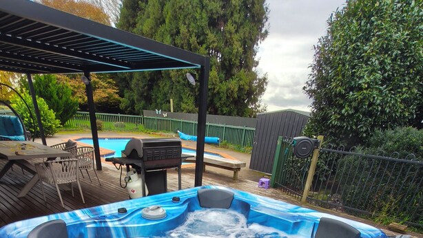 Un jacuzzi en una terraza de madera con una pérgola, rodeado por una piscina y mobiliario de exterior.