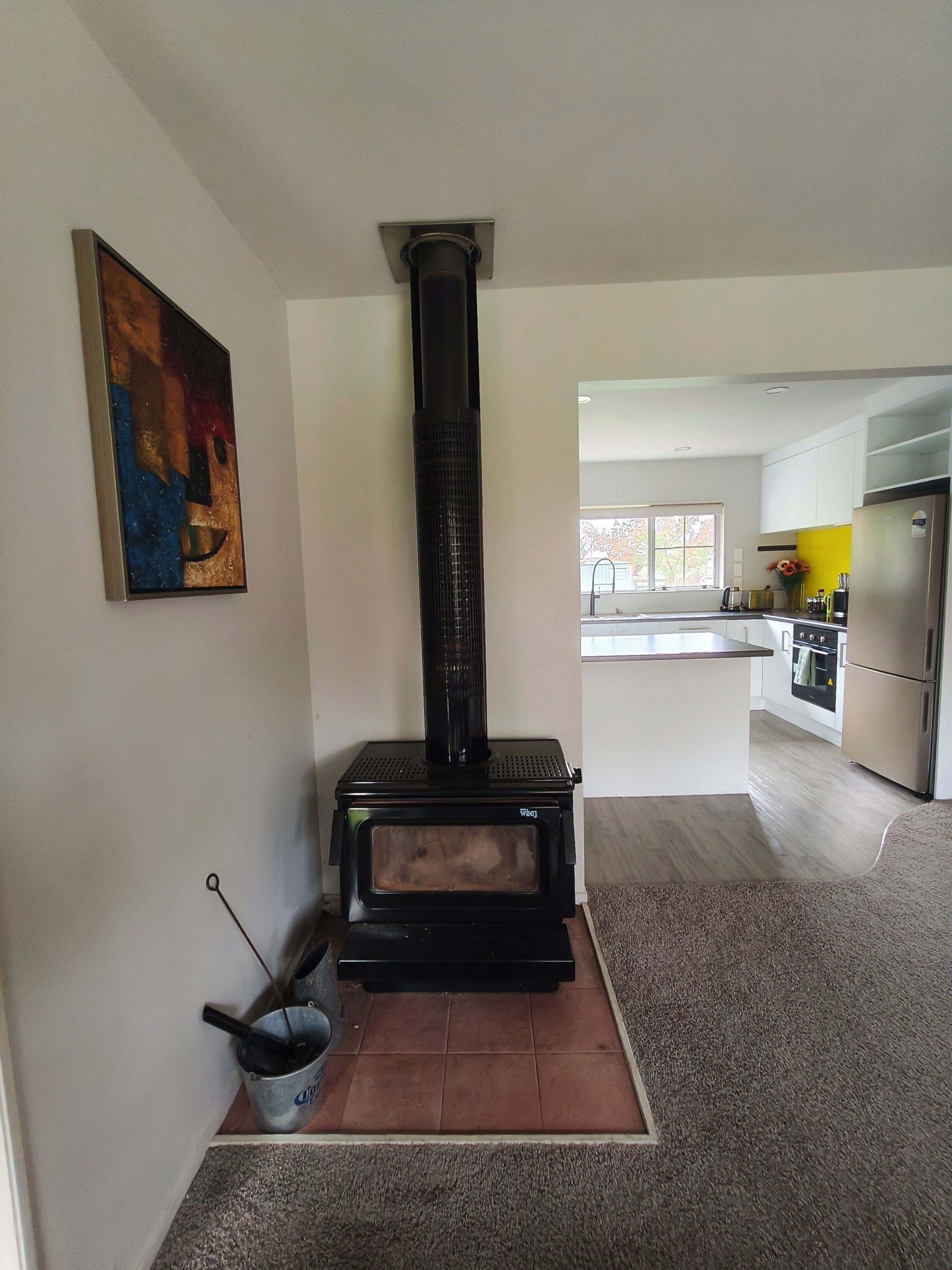 A modern kitchen with a built-in wood stove, a painting on the wall, and a view into the dining area.