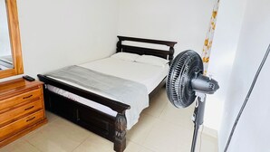 A small, single-bed room with a wooden dresser, a standing fan, and a ceiling vent.