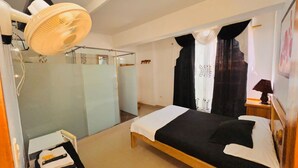 A bedroom with a bed, a bedside table with a lamp, a fan, a window with curtains, and a mirror.