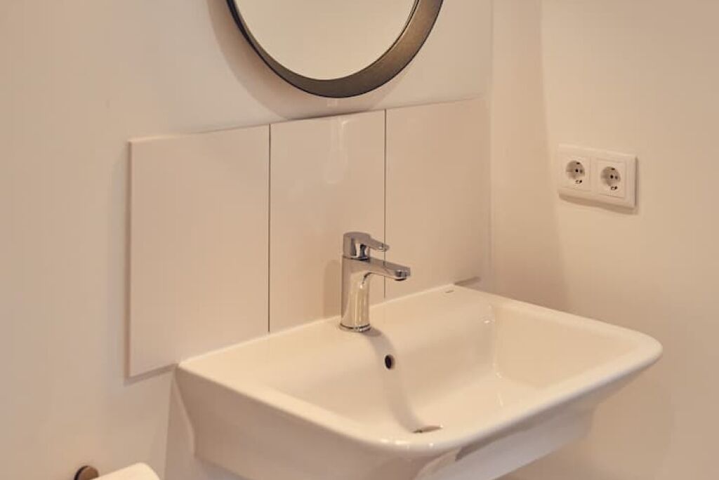 Un baño con un lavabo blanco, un espejo redondo y un enchufe en la pared.